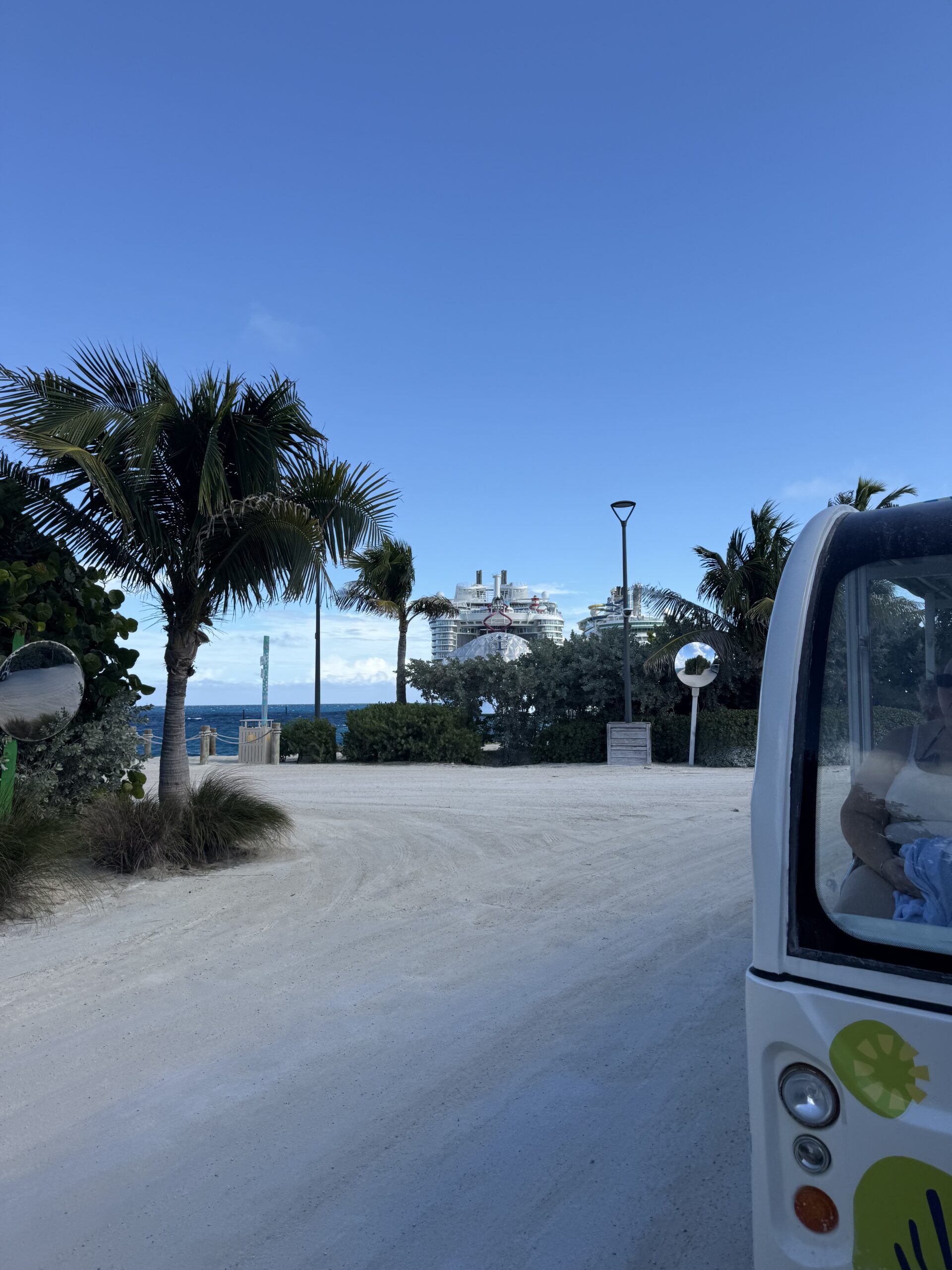 Sandy island road with palm trees, shuttle vehicle, and cruise ship visible in the distance at CocoCay