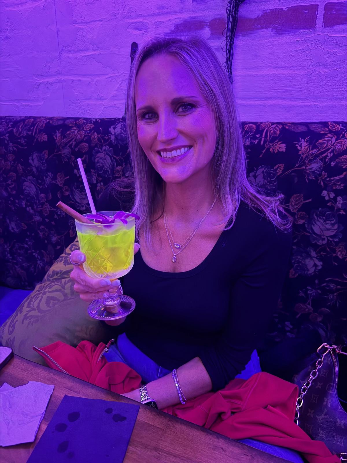 Danielle holding a yellow-green cocktail in the purple-lit seating area at BTW Spookeasy.