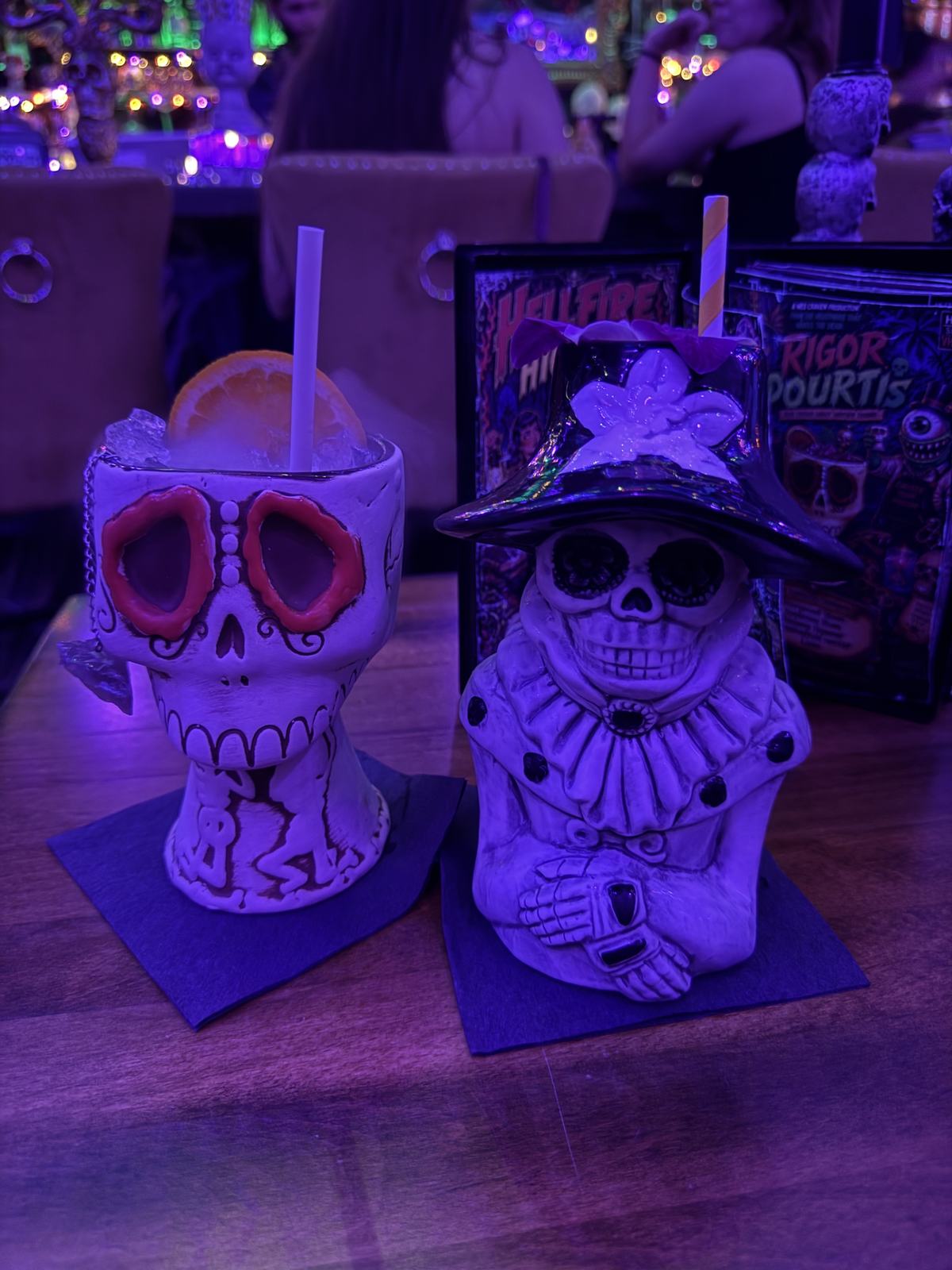 Two Halloween cocktails in skull-themed cups on the bar, with themed menu cards behind them.