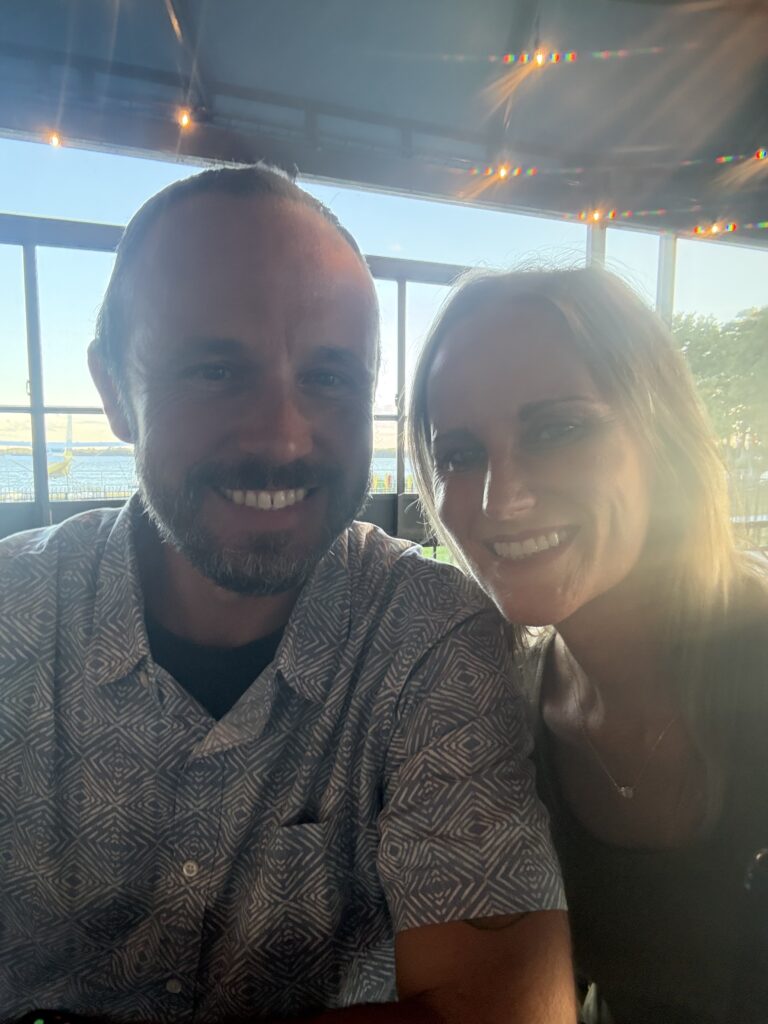 Smiling couple taking a selfie at Puddle Jumpers with the waterfront behind them