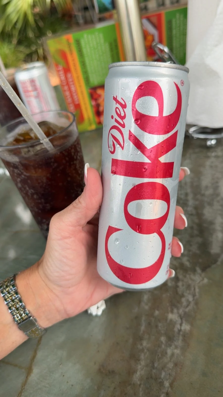 Hand holding a Diet Coke can beside a glass of soda and menus on an outdoor table at Tiki Bar & Grill.