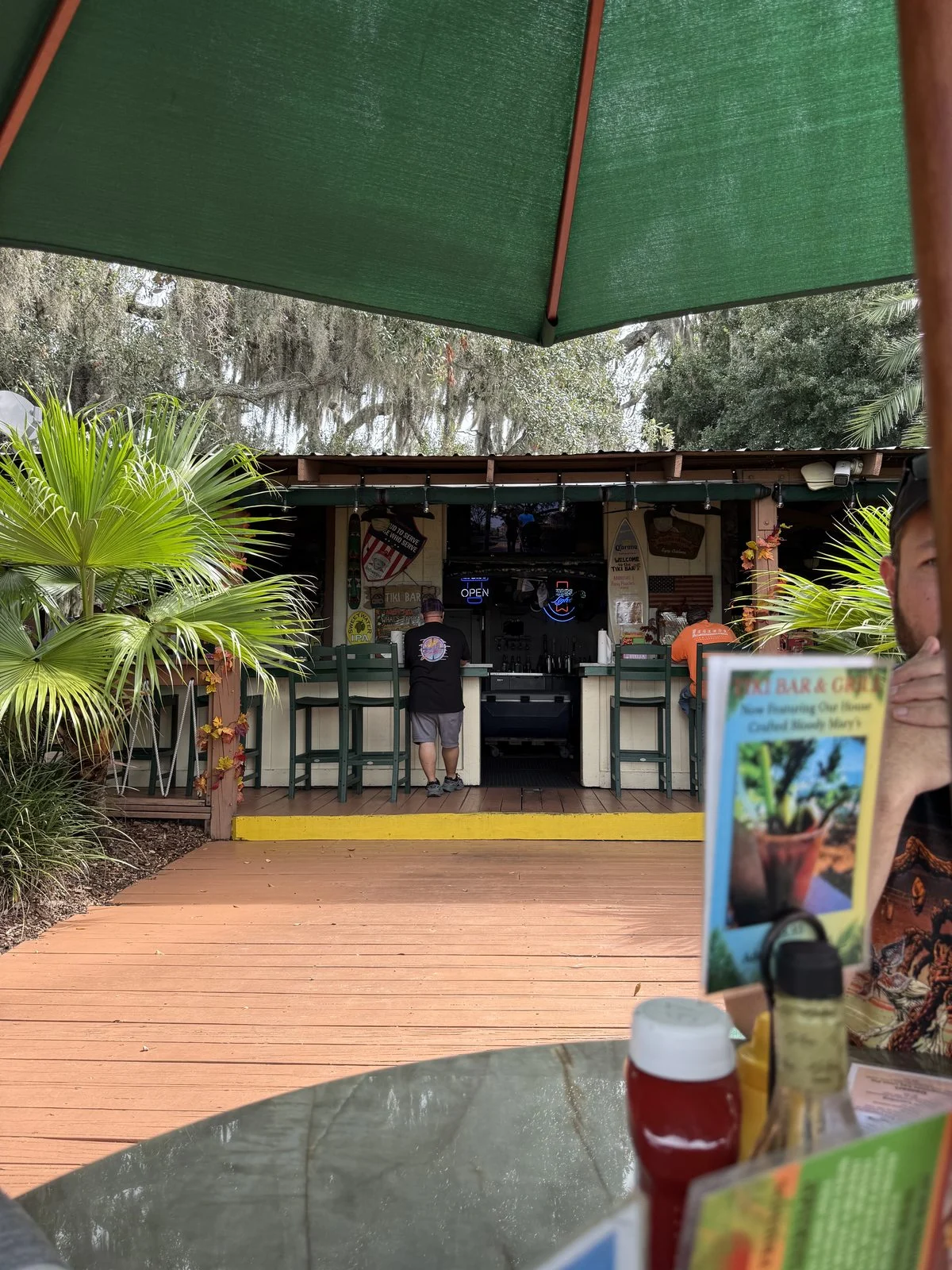Open-air tiki bar seen from an outdoor table beneath a green umbrella, with palm plants and Spanish moss nearby.