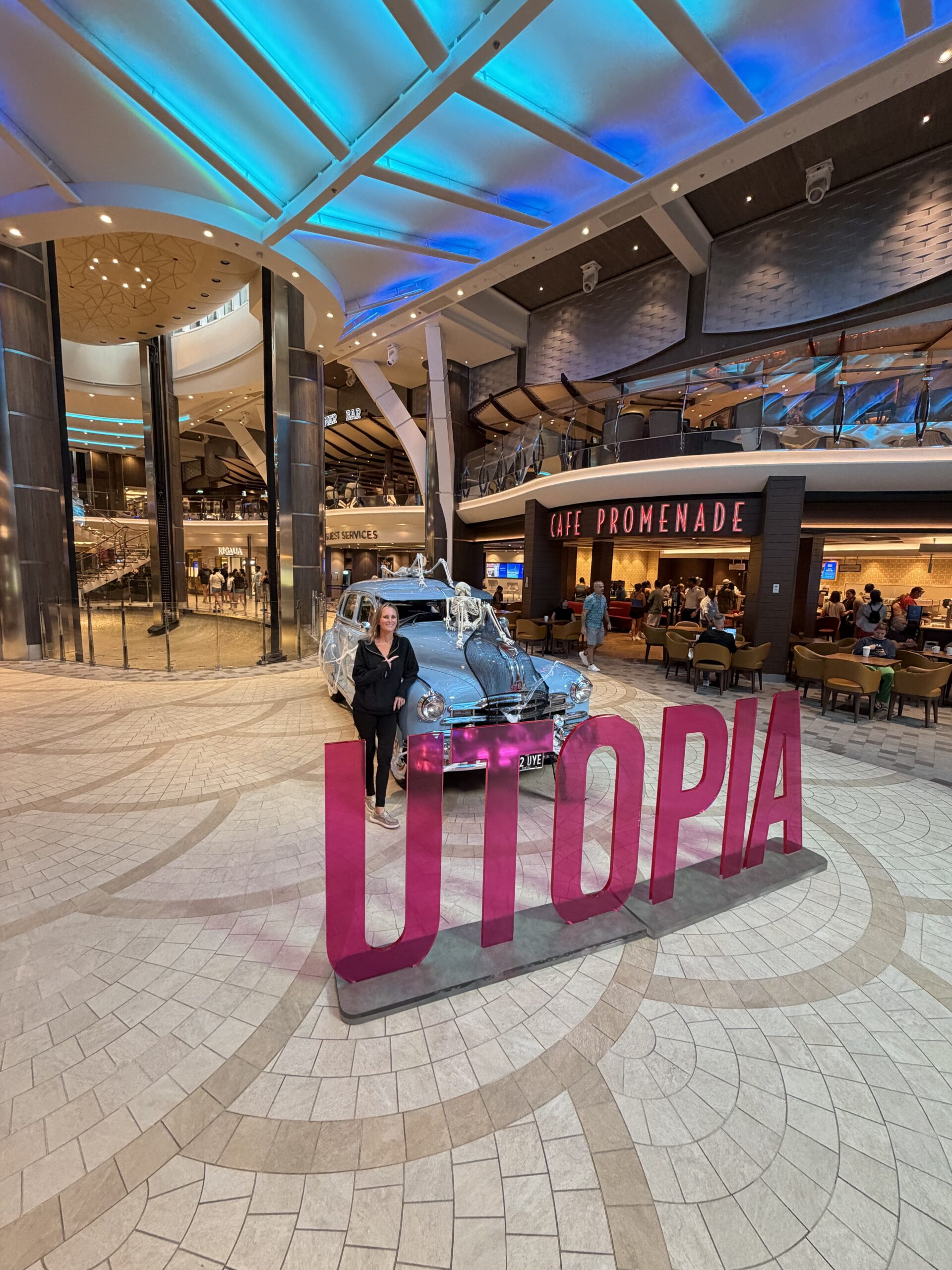 Woman standing beside a vintage car and pink UTOPIA sign on the Royal Promenade aboard Utopia of the Seas